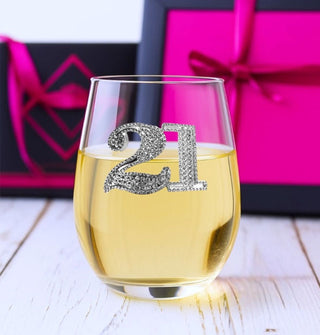 A stemless wine glass with silver jewel-encrusted "21" emblem is 2/3 filled with white wine against a backdrop of black and pink wrapped gifts