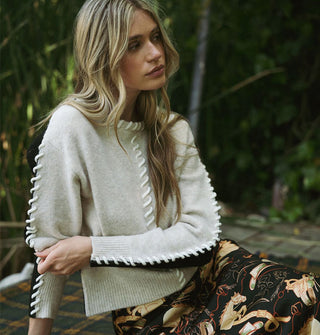 Model sitting in a forest setting wears a cream-colored sweater with black accent panels and white whip stitching down the arms, front center, and neckline