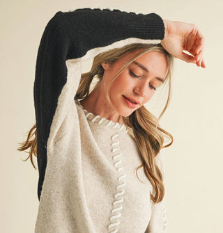 Model wears a cream-colored sweater with black arm panel and whip stitching along the neckline and down the front center