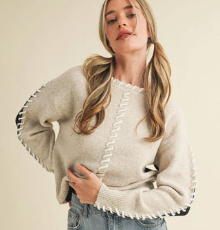 Model wears a cream-colored sweater with black arm panels and whip stitching along the neckline and down the arms and front center