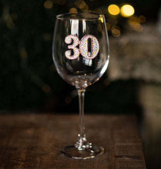 Clear wine glass on wooden tabletop features a central "30" insignia accented with colorful jewel embellishments