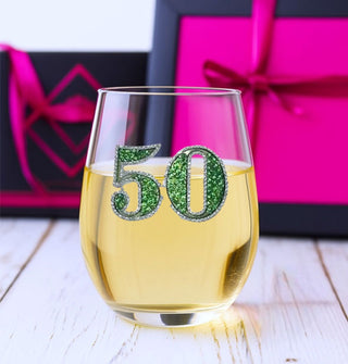 Rhinestone-encrusted "50" decal stemless wine glass is 2/3 filled with white wine and is set against a backdrop of black and pink wrapped presents