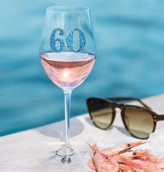 Bejeweled "60" wine glass sitting at the side of a pool with sunglasses and a plate of shrimp is half-filled with pink prosecco