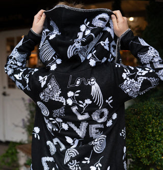 Model pulls up the hood of a black cardigan with white embroidered designs all over it