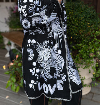 Model standing against a backdrop of potted plants on a porch deck wears a long black cardigan with white embroidered designs all over it