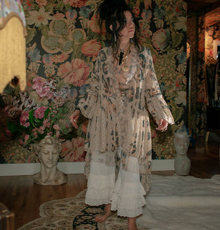 Model standing against a floral backdrop wears a lace open-front mid-length duster with enlarged leopard print and tiered white pantaloons