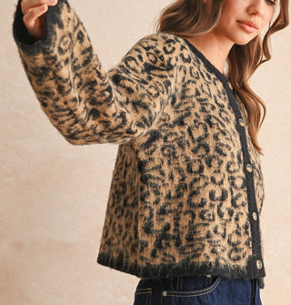 Model with arm upraised wears a leopard print button-down cardigan with black placket, neckline, and sleeve cuffs paired with a dark wash denim skirt