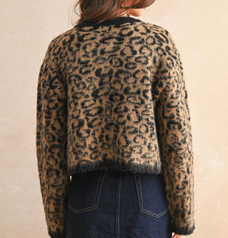 Model with back turned to camera wears a leopard print button-down cardigan with black placket, neckline, and sleeve cuffs paired with a dark wash denim skirt