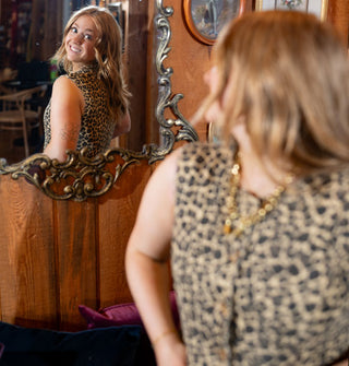 Model wearing a leopard print vest looks over her shoulder at her smiling reflection in an ornate, gold-framed mirror on the wall behind her