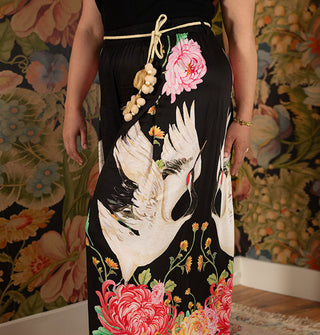 Model wears ultra wide-legged black pants with large print of flowers and cranes, accessorized with a tassel and pom belt