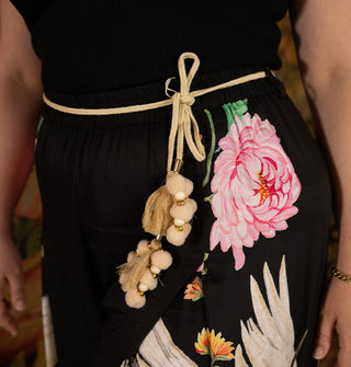 Model wears black pants with large floral and crane print accessorized with a tied  tassel and pom belt