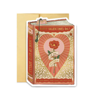 Greeting card resting in front of a gold envelope is designed as a book titled, "A Book of Love Spells" and says, "Happy Valentine's Day" at the top in gold lettering and features a central rose illustration inside a radiant heart with the message, "I am bewitched by your beauty, your spark, and your soul," also in gold lettering and partially written on a banner wrapped around the rose's stem