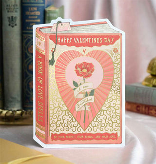 Greeting card is designed as a book titled, "A Book of Love Spells" and says, "Happy Valentine's Day" at the top in gold lettering and features a central rose illustration inside a radiant heart with the message, "I am bewitched by your beauty, your spark, and your soul," also in gold lettering and partially written on a banner wrapped around the rose's stem