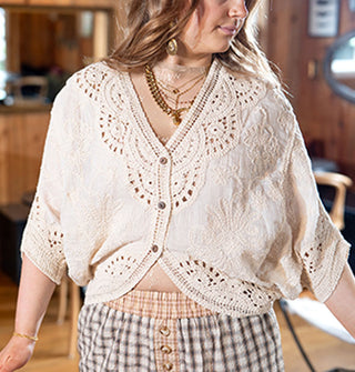 Model wears a white crocheted top with 3/4 dolman sleeves, three buttons at the front, and a deep V neckline paired with plaid pants and gold jewelry