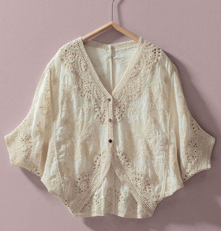 White crocheted top with 3/4 dolman sleeves, three buttons at the front, and a deep V neckline hangs on a clothes hanger against a light purple backdrop