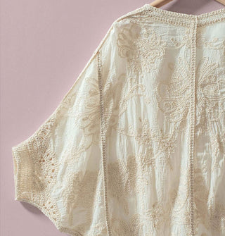 White crocheted top with 3/4 dolman sleeves hangs on a clothes hanger against a light purple backdrop