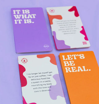 Sample MidZen affirmation cards, the purple and orange backs of which say, "It Is What It Is"m and "Let's Be Real" in white lettering