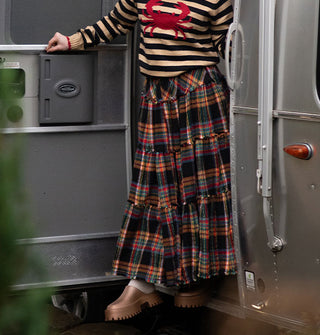 Model stepping out of an Airstream trailer wears a beige and black striped sweater with red crab decal paired with a multicolored plaid ruffle-tier skirt and copper clogs