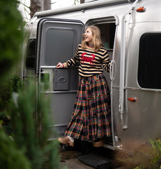 Model stepping out of an Airstream trailer wears a beige and black striped sweater with red crab decal paired with a multicolored plaid ruffle-tier skirt and copper clogs