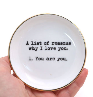 Model's hand holds a round white dish with gold rim and black stamped lettering that says, "A list of reasons why I love you. 1. You are you." 