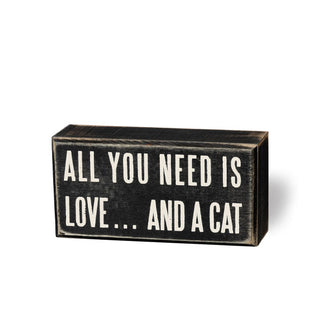 Rectangular black box sign with distressed finish says, "All you need is love... and a cat" in large white lettering