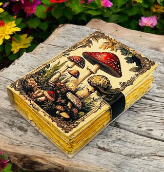 Notebook with antique-style mushroom illustrated cover, rough page edges, and closure strap with brass lock rests on a rustic wooden surface against a garden backdrop