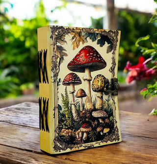 Mushroom notebook with black X spine binding and brass lock sits on end on a wooden surface against a garden backdrop