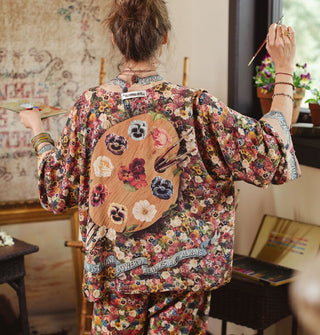 Model holding a paint palette and paintbrush and facing away from the camera wears a densely floral-patterned silken top with paint palette illustration on the back in which the paint daubs have been replaced by flowers