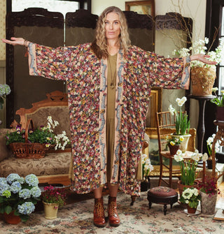Model standing with arms outstretched wears a densely floral patterned robe with contrasting trim in a room staged with antique furniture and flowers