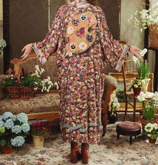 Model with arms outstretched facing away from the camera wears a densely floral-patterned robe with artist's palette design on the back in a room staged with antique furniture and flowers
