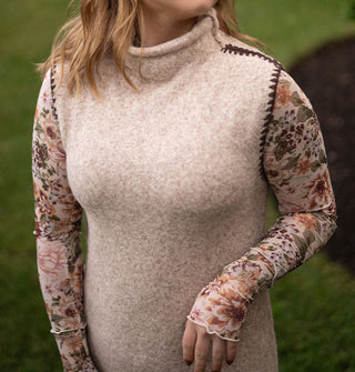 Model wears a sleeveless beige turtleneck sweater dress with brown stitching around the armholes paired with a floral long-sleeved mesh top