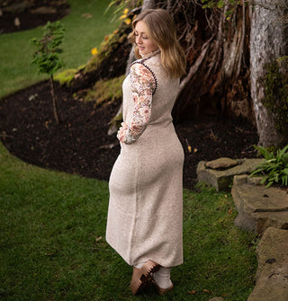 Model wears a maxi-length sleeveless beige turtleneck sweater dress with brown stitching on the armhole and shoulder paired with a floral long-sleeved mesh top
