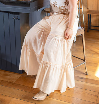 Model sitting on a bar stool wears light peachy-pink, wide-legged pants with horizontal ruffled tiers