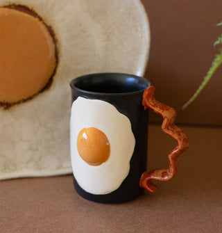 Black coffee mug features a fried egg design on its body and a handle designed to resemble a strip of bacon