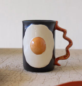 Black coffee mug features a fried egg design on its body and a handle designed to resemble a strip of bacon