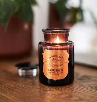 A lit Apothecary Noir Paddywax Baltic Ember candle shines from inside a black apothecary-style jar on a wooden surface with houseplants in the background