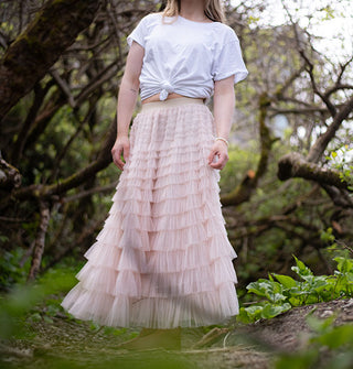 Model wears a multi-tiered ruffled light pink tulle maxi skirt with knotted white T-shirt in a forest setting