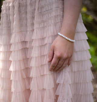 Model's hand with white beaded bracelet on wrist falls against a multi-tiered ruffled light pink tulle skirt in a sun-dapped forest setting