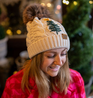 Smiling model wears a white cable-knit beanie with green tree embroidery and faux fur pompom on top