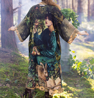 Model poses with arms outstretched in a misty wooded area wearing a highly decorative loose-fitting robe with woman's portrait prominently featured