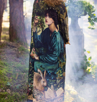 Model standing in a misty wooded area wears a highly decorative loose-fitting robe with woman's portrait prominently featured
