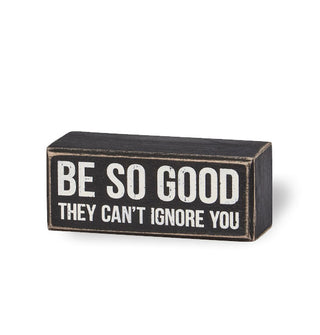 Rectangular black box sign with distressed finish says, "Be so good they can't ignore you" in large white lettering