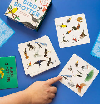 Model's hand points to a bird on one card from the Bird Spotter game, on a blue surface with other cards in the deck, the game box, and the green instruction booklet