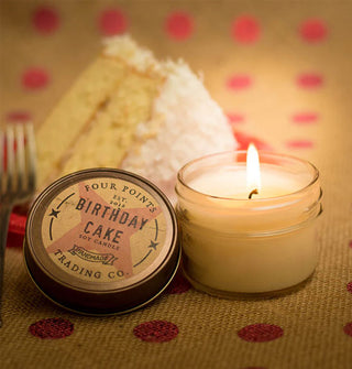 Lit Birthday Cake jar candle with lid removed is staged on a polka dot surface with a slice of cake in the background
