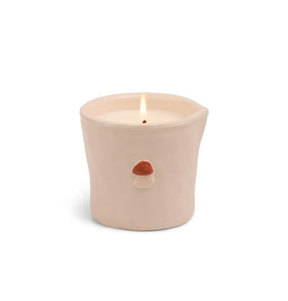 Irregular light pink ceramic candle vessel with lit wick features a small red and white raised painted mushroom insignia on the side