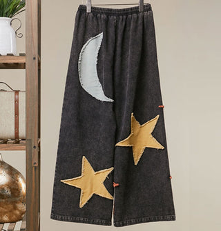 Black wide-legged pants with gray moon and yellow stars appliqués on the back, all with rough edges