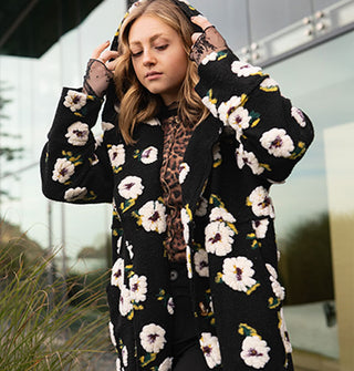 Model wears a black hooded coat with plush white floral pattern in front of a glass structure