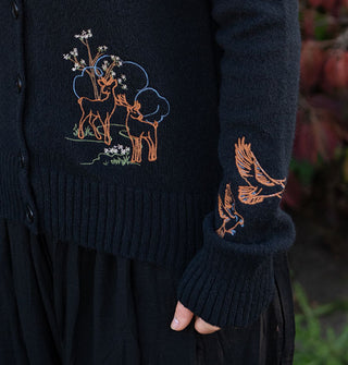 Model wears a black long-sleeved button-down cardigan with colorful embroidery of deer among plants, trees, and clouds on its bodice, and two birds in flight on its sleeve