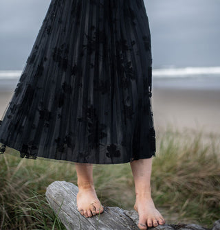 Barefoot model balancing on a piece of driftwood against a beach backdrop wears a black pleated floral tulle midi skirt