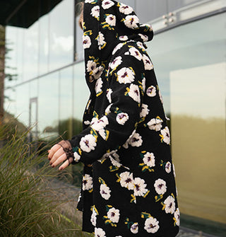Model with arm partially outstretched wears a black hooded coat with plush white floral pattern in front of a glass structure